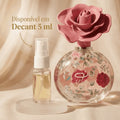 Perfume floral em frasco redondo com estampa de rosas e tampa em formato de flor rosa, ao lado de decant de 5ml sobre tecido rosa