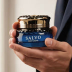 Creme corporal Salvo em pote de vidro azul com tampa dourada e detalhe canelado, segurado na mão.