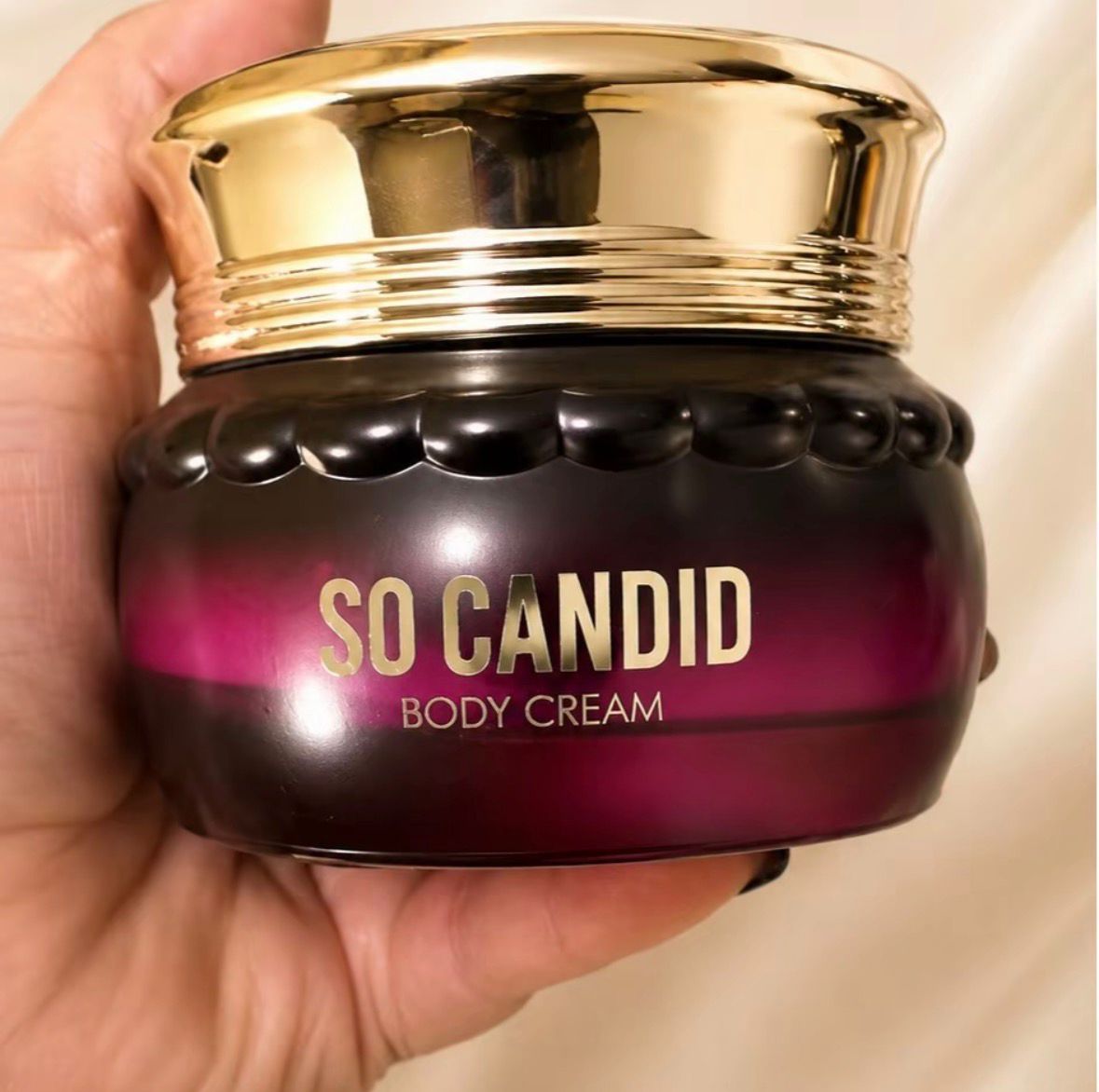 Creme corporal So Candid em pote de vidro vermelho com tampa dourada, segurado na mão.
